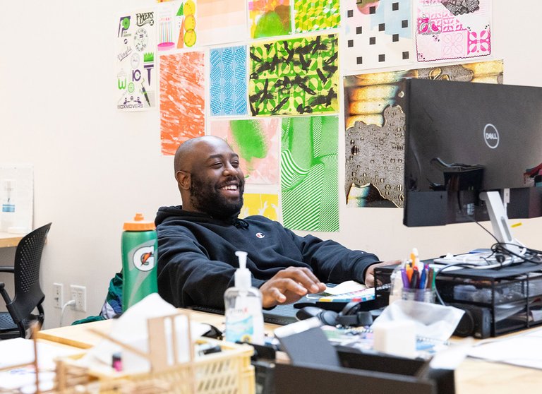 Malik Sapp in the MFA Design program smiles while he works. Behind Malik are various design patterns in different colors.