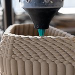 A large pencil-shaped apparatus 3D prints a small undulating structure with clay.