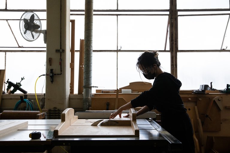 A student cuts wood wearing goggles and a face mask in a light-filled workshop.