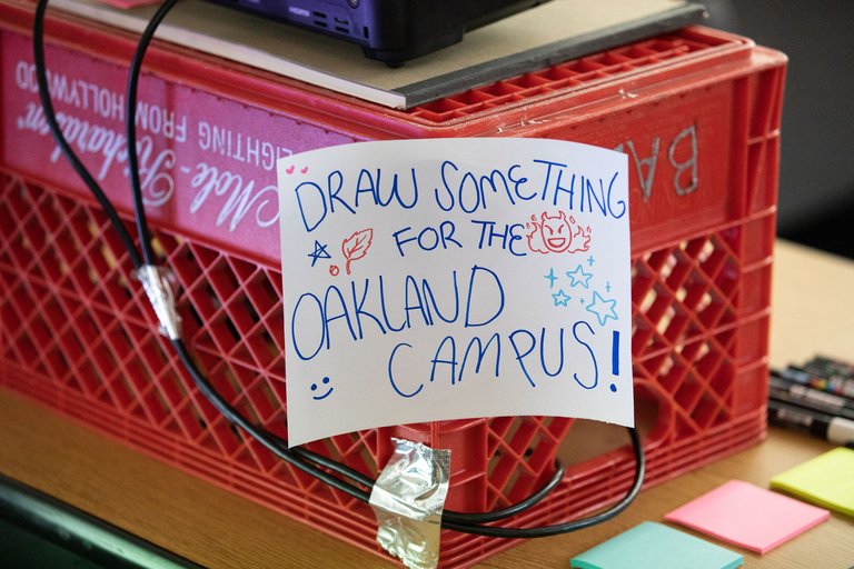 A photo of a hand-drawn sign on a red carton that says, “Draw something for the Oakland campus!”