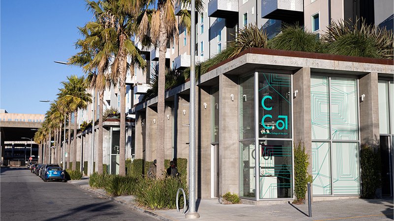 View from the street looking toward the corner of a modern-style building with the CCA logo in teal in the window. A row of palm trees lines the sunny street.