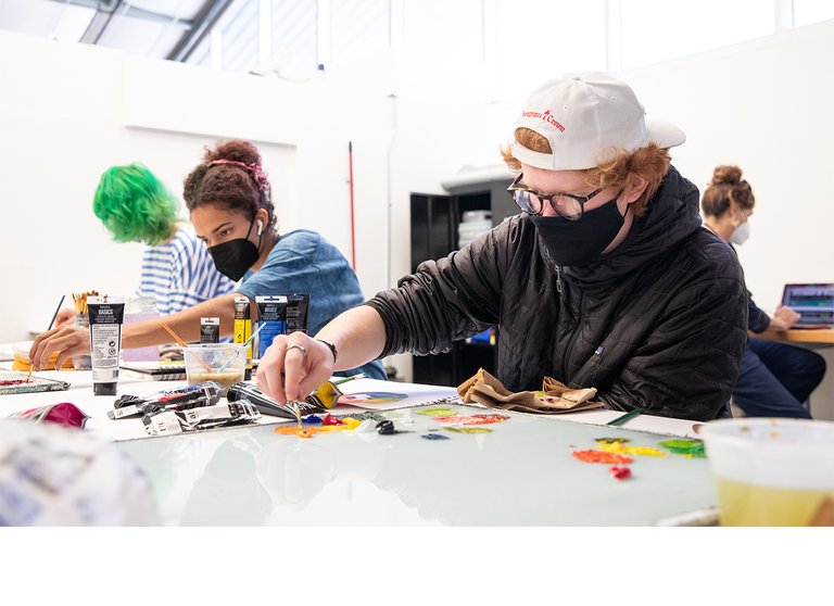 Three first-year CCA students seated at a work table mix colorful paint on glass palettes in a bright white art studio with natural light and vaulted ceilings.