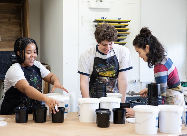 Three students work with art materials around a workbench.