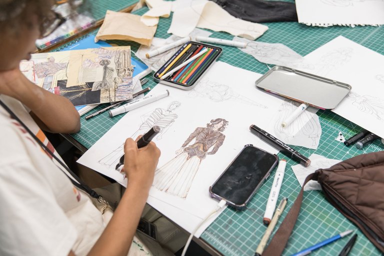 Person sketching fashion designs on paper at a workspace with colored pencils, markers, fabric swatches, and a smartphone nearby.