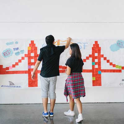 Two student work together to put up a poster of the Golden Gate Bridge made out of orange Post-It notes.