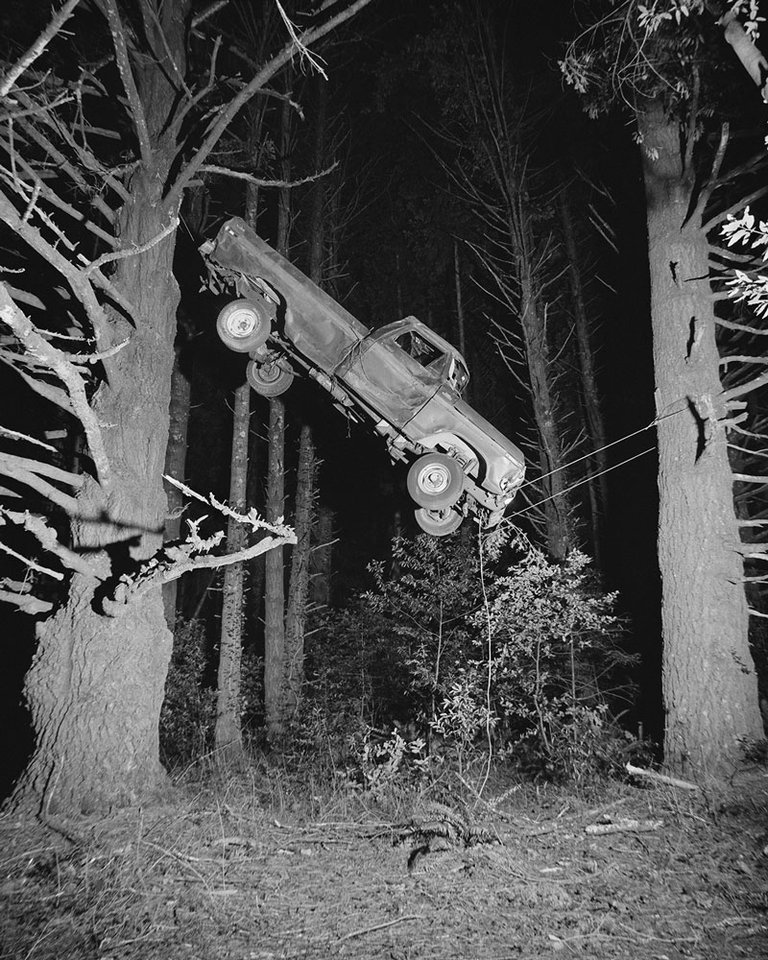 A black and white photograph of a car hanging from two trees.