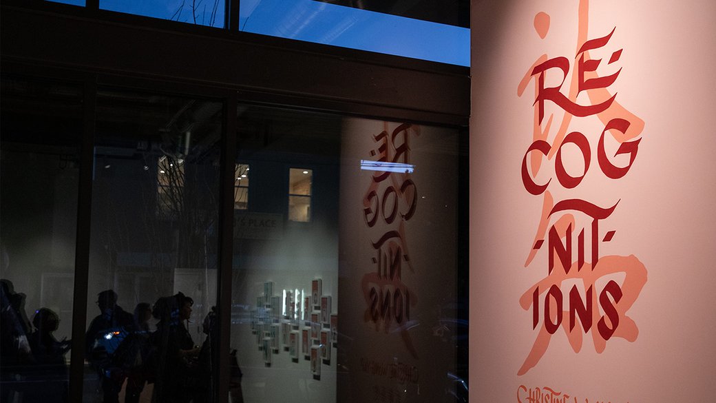 A peach colored wall with the word Recognitions painted on it is reflected in a window to the left.