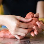 Close up image of a students hands sculpting red clay into a shape.