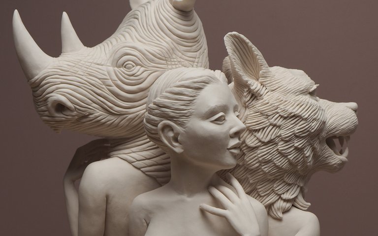 Crystal Morey, Three Graces, 2019. Hand-sculpted porcelain. A cropped image of three magical-realist monochromatic figures rendered in a smooth relief, two with animal heads—a rhino and a wolf. In the center, a head with upswept hair looks right. The central figure and rhino-head figure have a human hand in the frame, each touching their own collarbones.
