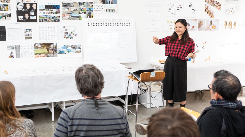 A woman in plaid gives a presentation amid a backdrop of different design artwork.