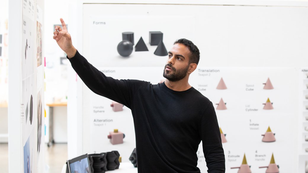 A man wearing black points to designs, with cone and cube shaped graphics behind him.