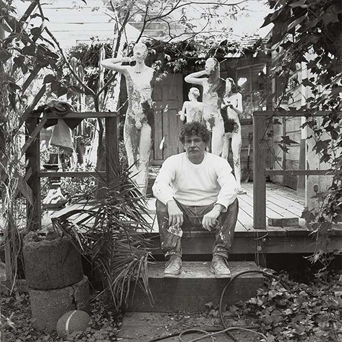 Manuel Neri sits in front of his sculptures in a black and white photograph