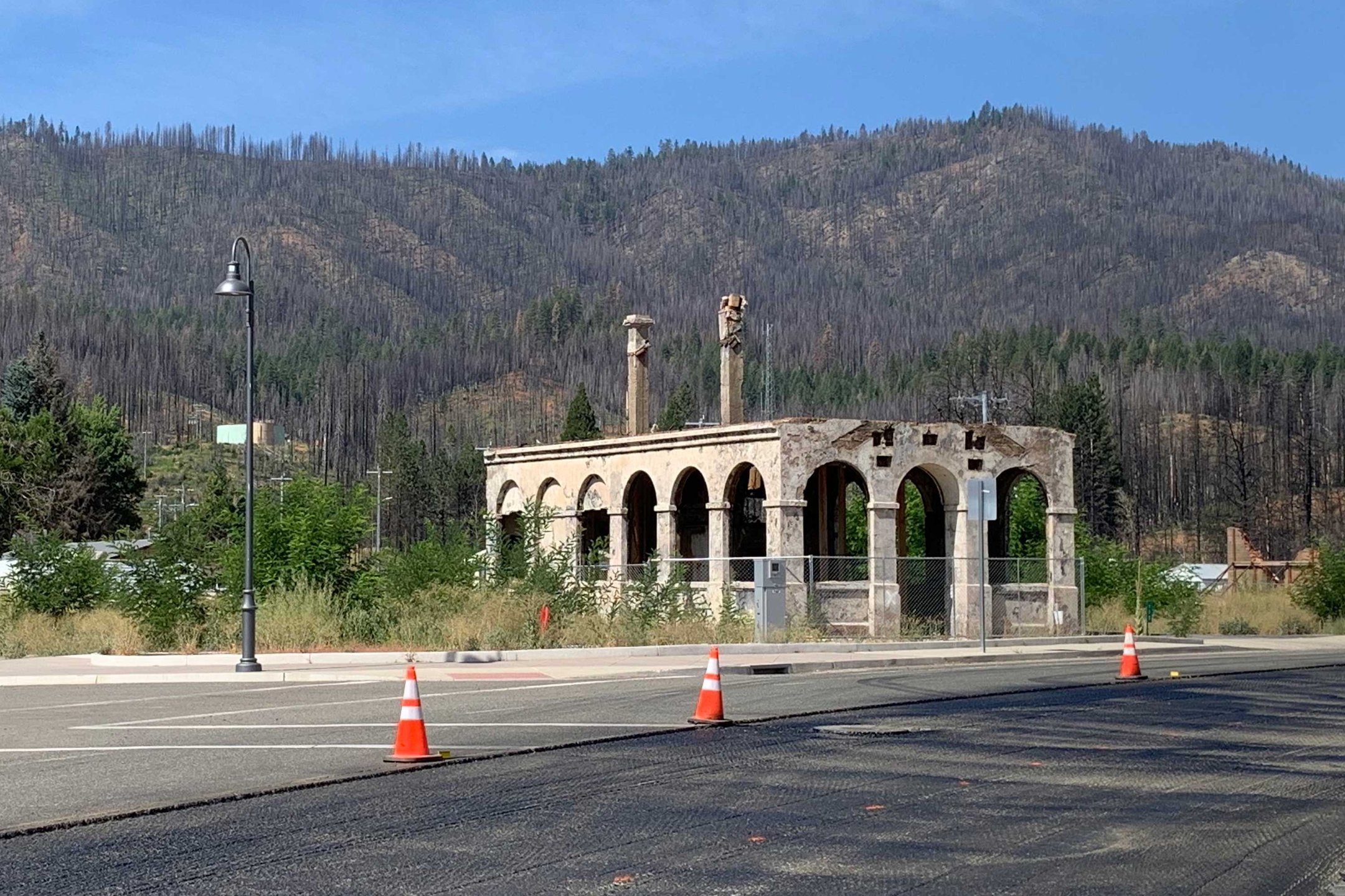 A photo of the remains of a single burned-out building amid a backdrop of forest regrowth and burned trees.