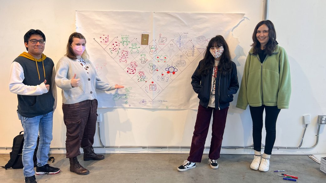 A group of four students posing in front of a large poster displaying a drawn grid with small characters in each box.
