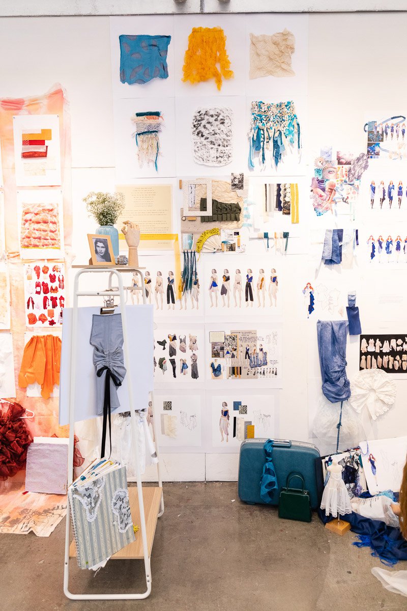 Fashion sketches and illustrations hang on a white wall, and fashion design-related objects sit on the concrete foreground.