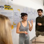 A woman with dark hair and a black-and-white striped jumpsuit talks about her project to a small group of people.