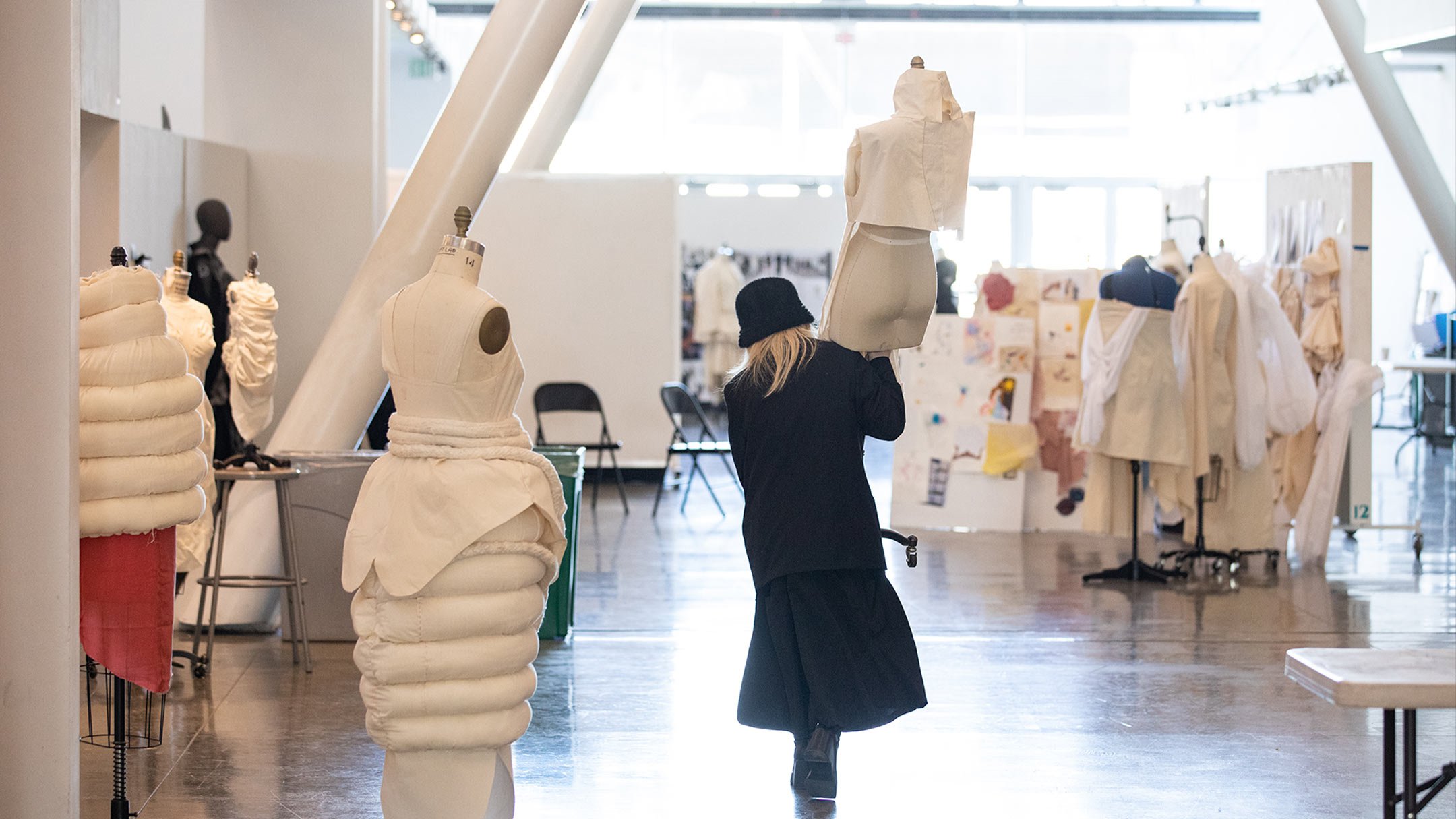A fashion design student dressed in black carries a dressmakers’ form through a well-lit space, surrounded by fashion sketches pinned to the wall and several more dress forms draped in white muslin garments.