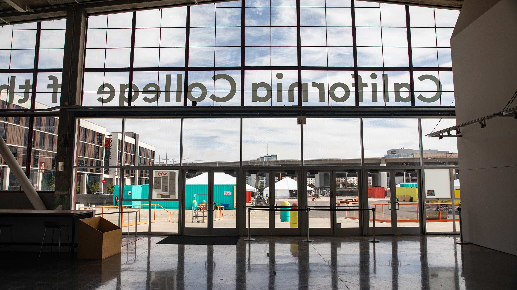 The Nave, an open, industrial space with large windows looking out to a sunlit asphalt lot dotted with white canopies and colorful shipping containers.