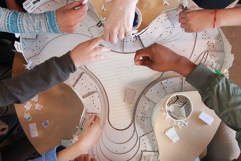 Seven students play a board game, arms reaching across a table with game pieces.
