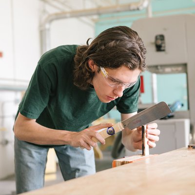 A student cuts wood wearing goggles and a face mask in a light-filled workshop.