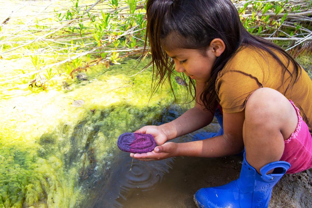 A photo of a young girl in blue boots holding a purple clay object over a small pond.