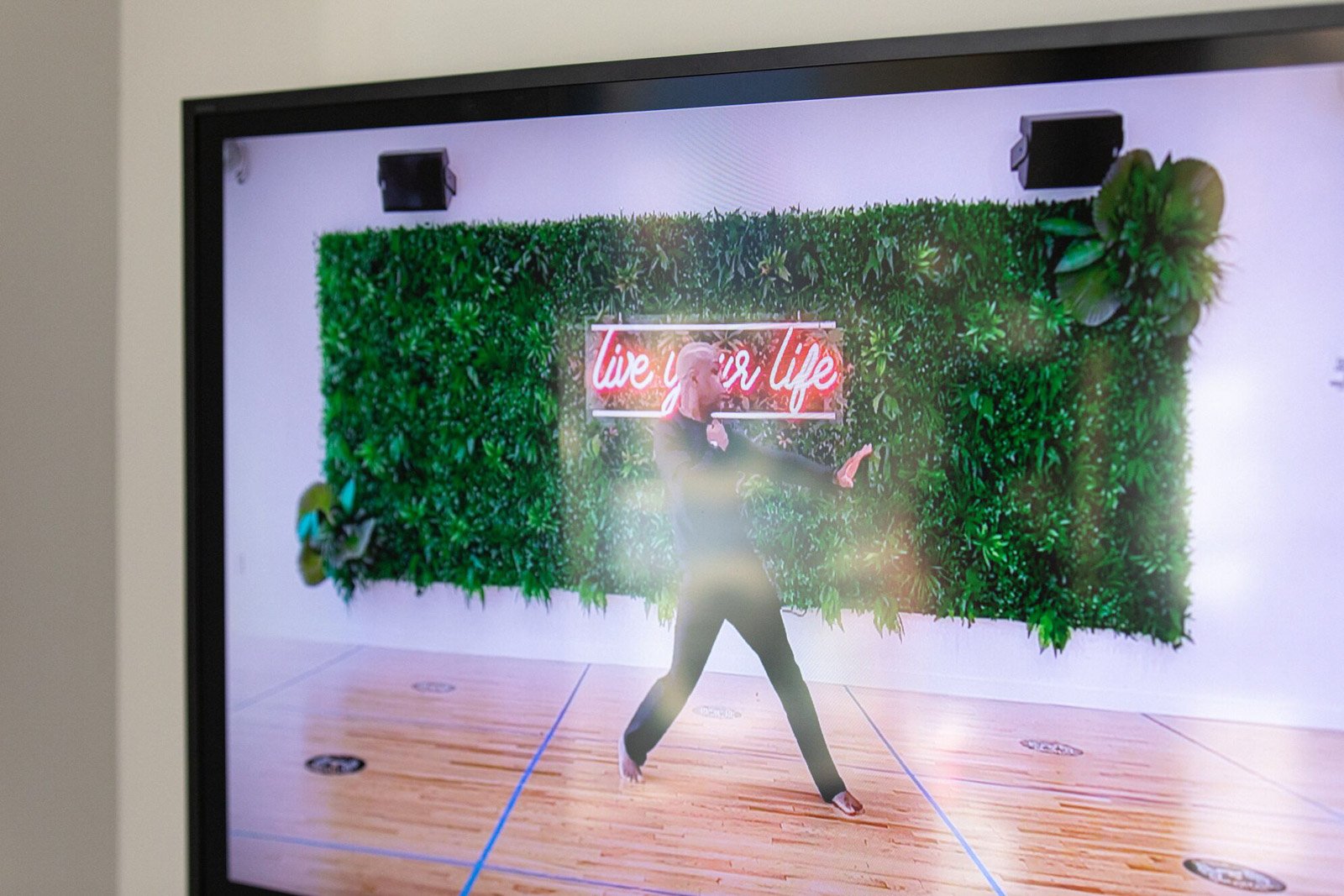 A video still of the artist dancing in front of a green wall with a neon sign that says live your life.