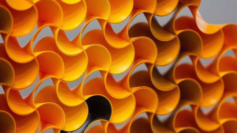 A macro photograph of curved orange paper forms creating an abstract geometric pattern. The paper is arranged in a repeating wave-like structure, with each curve flowing into the next to create a honeycomb-like effect.
