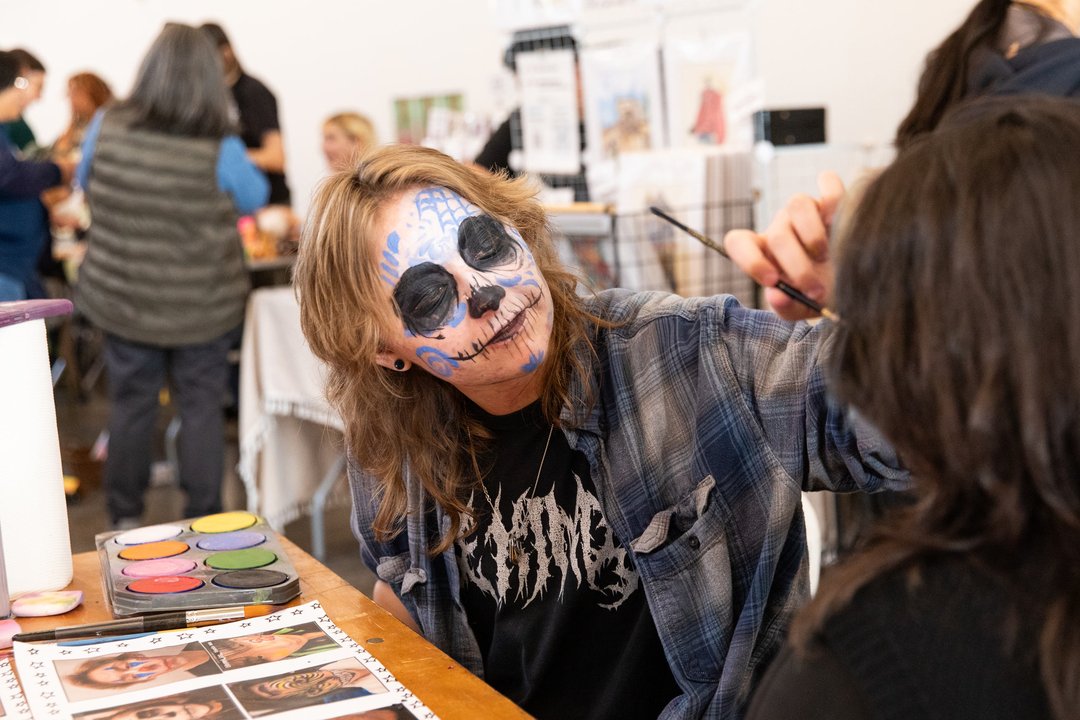 Person with blue and white skull face paint applies makeup to another person indoors, with face paint supplies and reference images on the table.