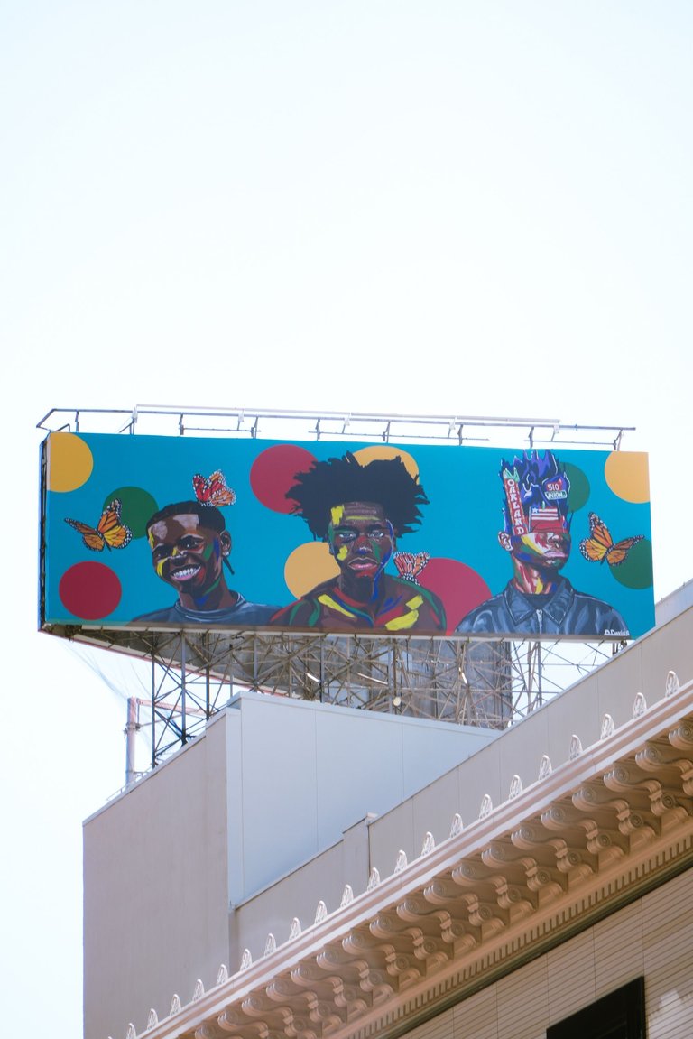 A colorful billboard featuring illustrated portraits with vibrant circular patterns is mounted on a metal frame above a beige building, with a traffic light visible in the foreground against a bright turquoise sky.