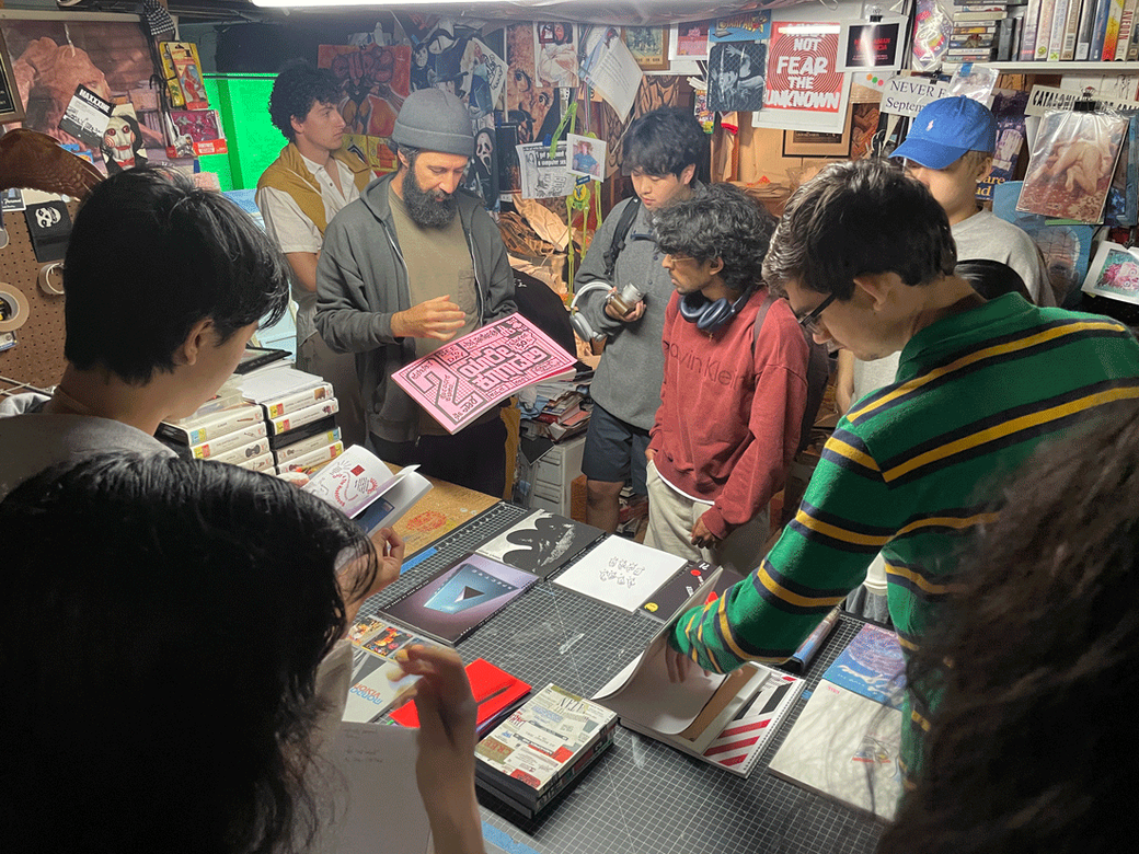 People gathered around a table covered with books, zines, and printed materials, with someone in a green and yellow striped sweater examining items on a cutting mat.