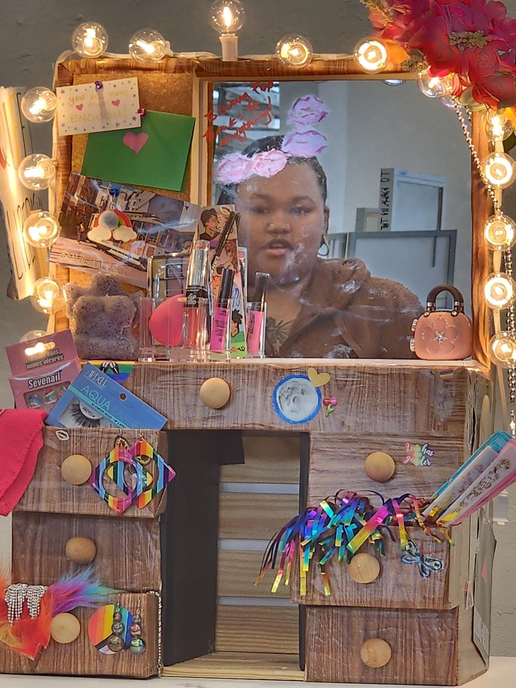 A photo of a handmade vanity with theatrical lights, rainbows, LGBT ephemera, and a mirror reflecting the artist.