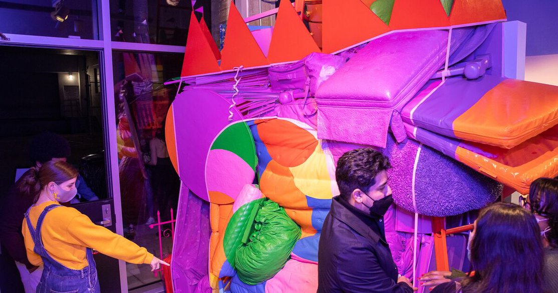 Visitors look at a monumental sculptural artwork comprised of various pieces of found furniture and painted in bright purple, orange, and green.