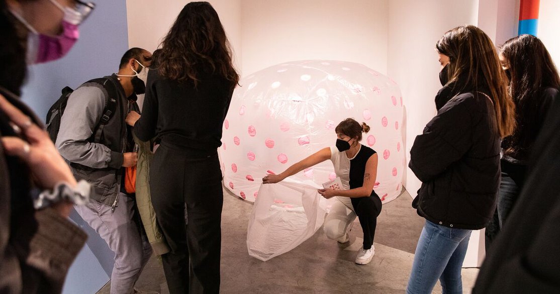 A student holds open a small entrance to an inflated plastic sculpture painted with pink polka dots.