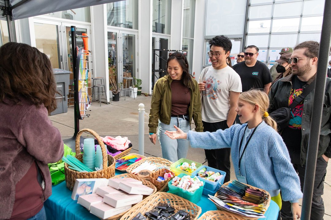 People gather around a table displaying various items, engaged in conversation and examining the objects.