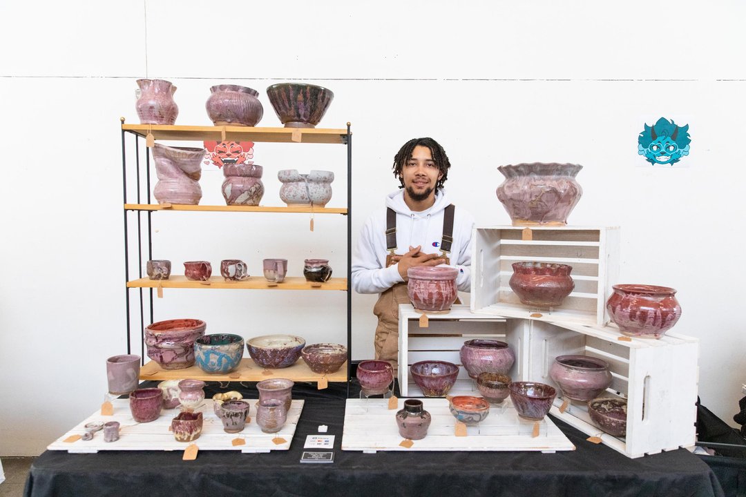 A student stands behind a display of handmade ceramics arranged on shelves and crates at a booth.