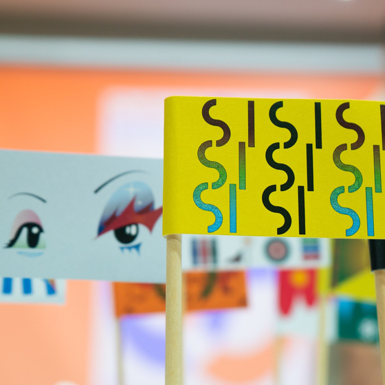 Close-up of handmade paper flags on sticks—one yellow with black squiggles, one mint with illustrated eyes—in a colorful gallery.