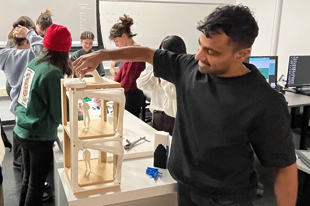 A smiling man in a black shirt interacts with a wooden model at a classroom table. Other students work collaboratively in the background.