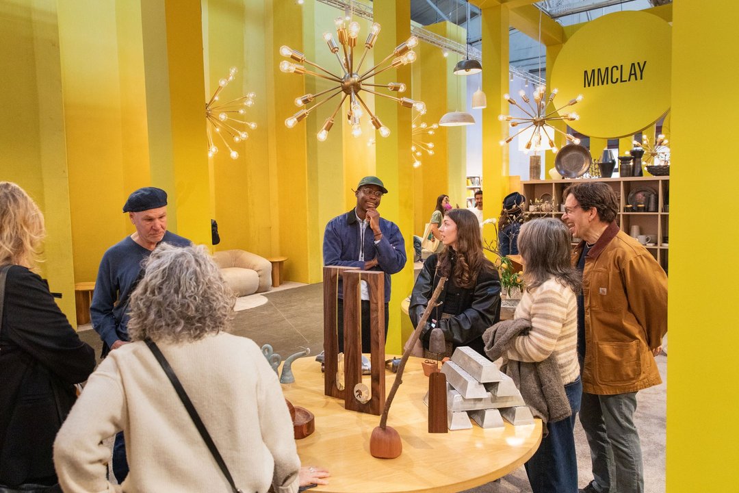 This editorial image depicts a vibrant, modern showroom space filled with a group of people engaged in conversation. The room is bathed in a warm, golden glow from the dramatic, sputnik-style chandeliers overhead, creating a cozy and inviting atmosphere.  The people in the image appear to be a mix of customers and staff, with several individuals standing and conversing in small groups. The group includes a diverse range of ages, genders, and ethnicities, suggesting a welcoming and inclusive environment.