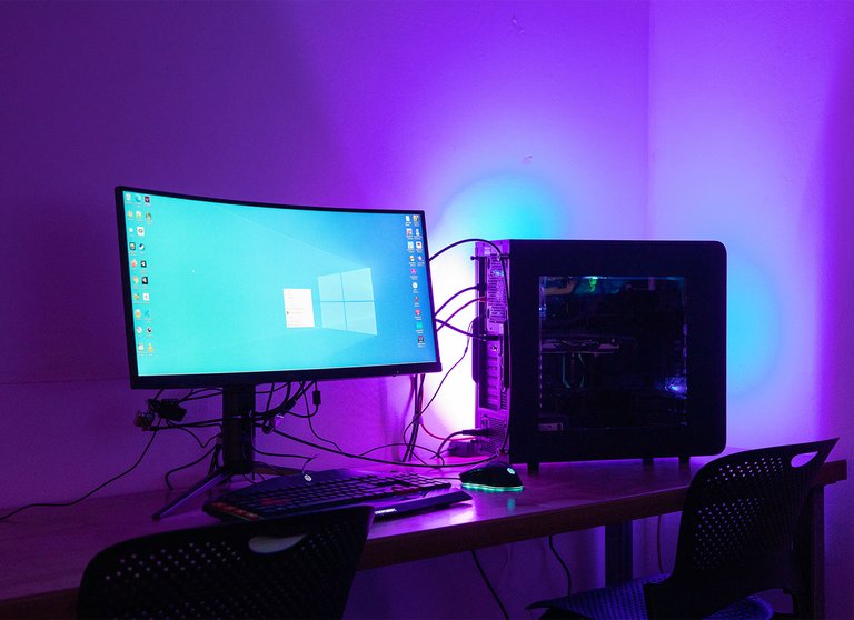 A desktop computer, monitor, and mouse are illuminated in purple and green hues in a dark room.