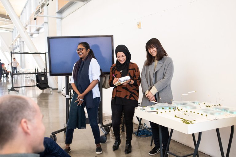 Three students laugh next to their architectural model during a critique.