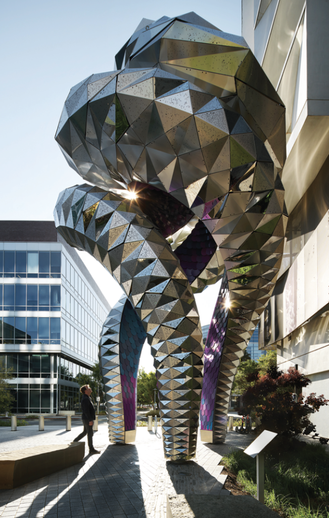 A tall, highly reflective serpentine structure that bounces sunlight from its geometric, shiny, stainless-steel tiles.