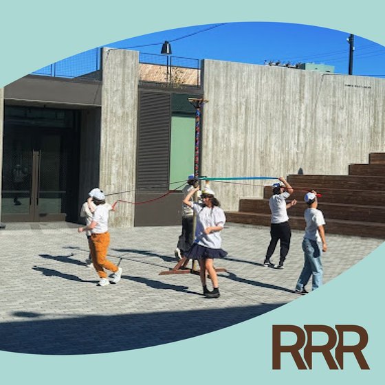 This editorial image depicts a group of people engaged in an outdoor activity or performance on a concrete plaza. The setting features a modern, minimalist architectural style with large, textured concrete walls and a bright blue sky in the background.   The people in the image appear to be young adults, dressed casually in t-shirts, jeans, and sneakers. They are positioned in various poses, some standing, some crouching, and some interacting with each other.