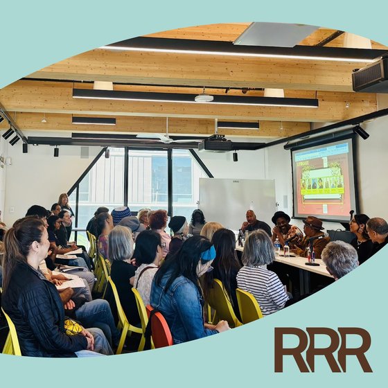 This image depicts a crowded indoor event or presentation, likely a conference or panel discussion. The room has a modern, open-concept design with exposed wooden beams and track lighting overhead. A large projection screen is visible at the front of the room, suggesting a presentation or speaker is taking place.  The audience is composed of a diverse group of people, including both men and women of varying ages and backgrounds.