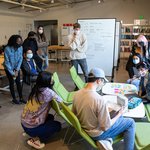 A group of twelve students gather in a circle around a table with post-its.