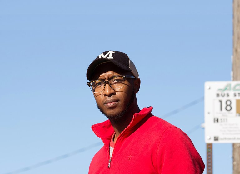 A portrait of Shane T. Watson with a bright blue sky in the background. He is wearing a red sweater and a hat with an M on it. Part of a bus stop sign can be seen behind him.