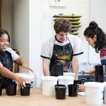 Three students work with art materials around a workbench.