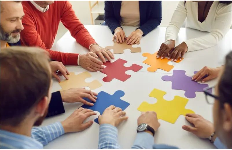 Diverse group collaborating around a white table, assembling colorful puzzle pieces to symbolize teamwork
