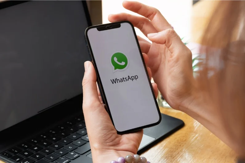 Person holding a smartphone with WhatsApp logo, laptop in background on wooden desk, symbolizing digital communication.
