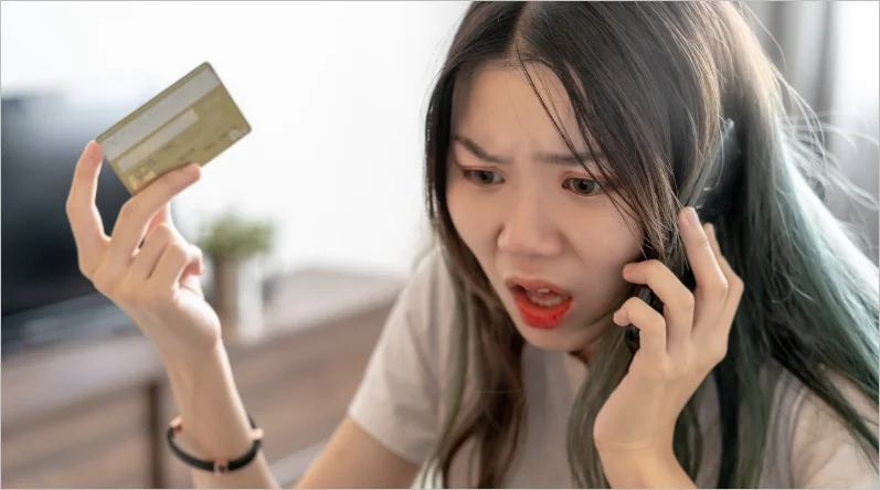 Woman on phone holding a credit card, looking shocked and concerned, suggesting an issue with her card.