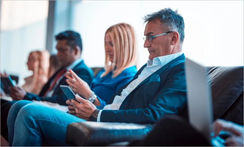 Group of people seated in a row, focused on smartphones and tablets in a professional, calm setting.